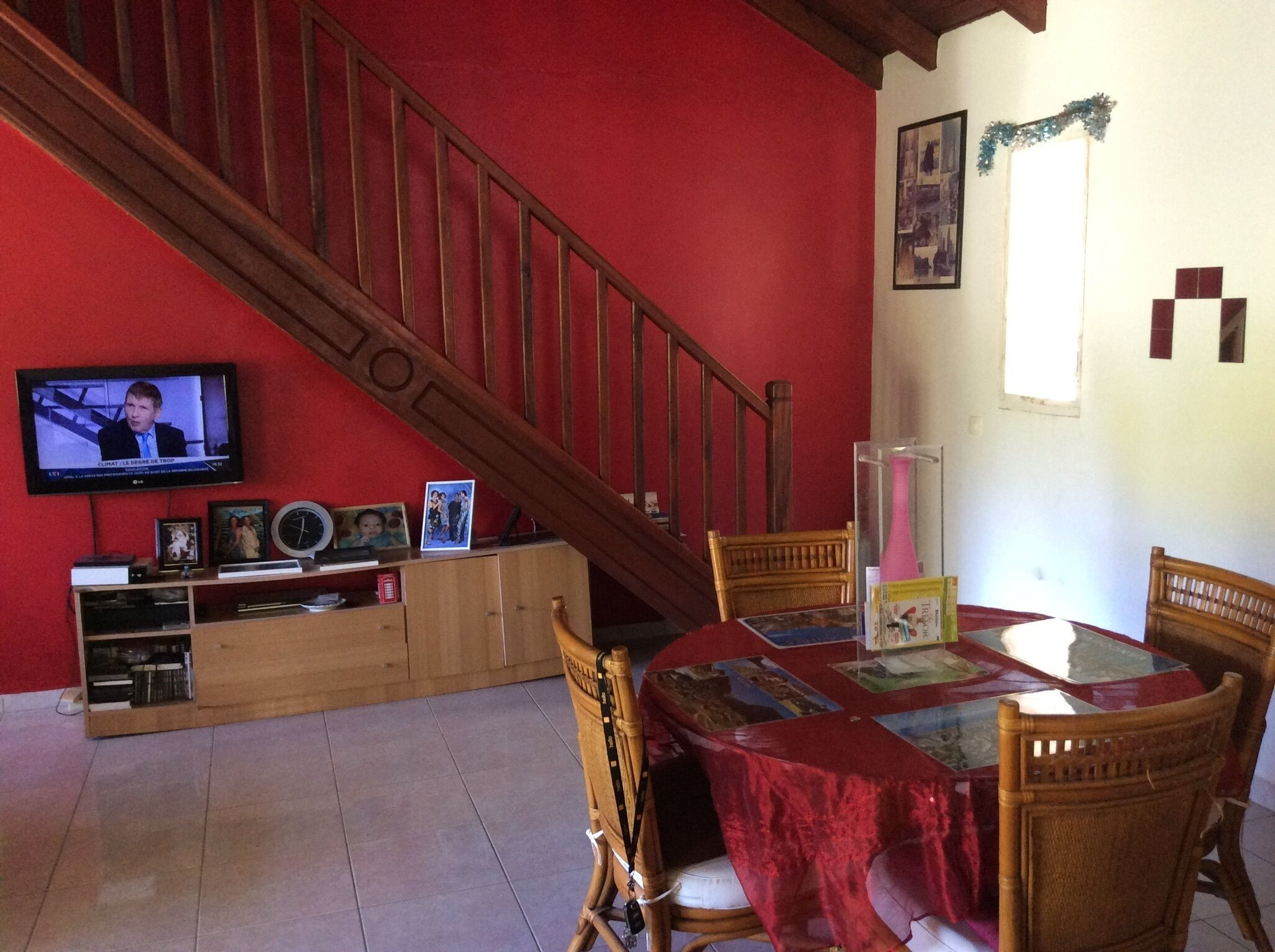 A dining area with a wooden table covered in a red tablecloth, surrounded by wicker chairs. A television is mounted on the wall above the table, displaying a news program.