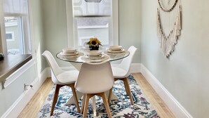 A dining area with a round glass table, white chairs, a hanging pendant light, and a macrame wall hanging. - Beautiful Victorian with Modern Flare close to Town, Train & Beach (Ipswich)
