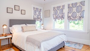 A bedroom with a large bed, two bedside tables, a nightstand, a window with patterned curtains, and a wooden floor. - Beautiful Victorian with Modern Flare close to Town, Train & Beach (Ipswich)
