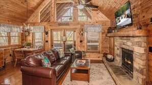 A log cabin interior with a fireplace, a leather sofa, a dining area, and a television mounted on the wall. - Cozy Cabin w/ Private Fishing Dock & Hot Tub (Sevierville)