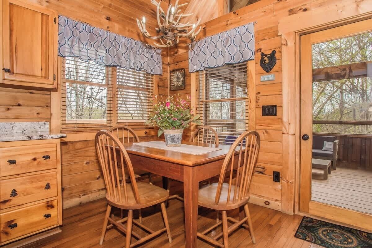 Interno di una baita in legno con tavolo, sedie, un lampadario e vista sull'esterno.