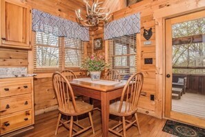 A wooden cabin interior with a dining table, chairs, a chandelier, and a view of the outdoors.