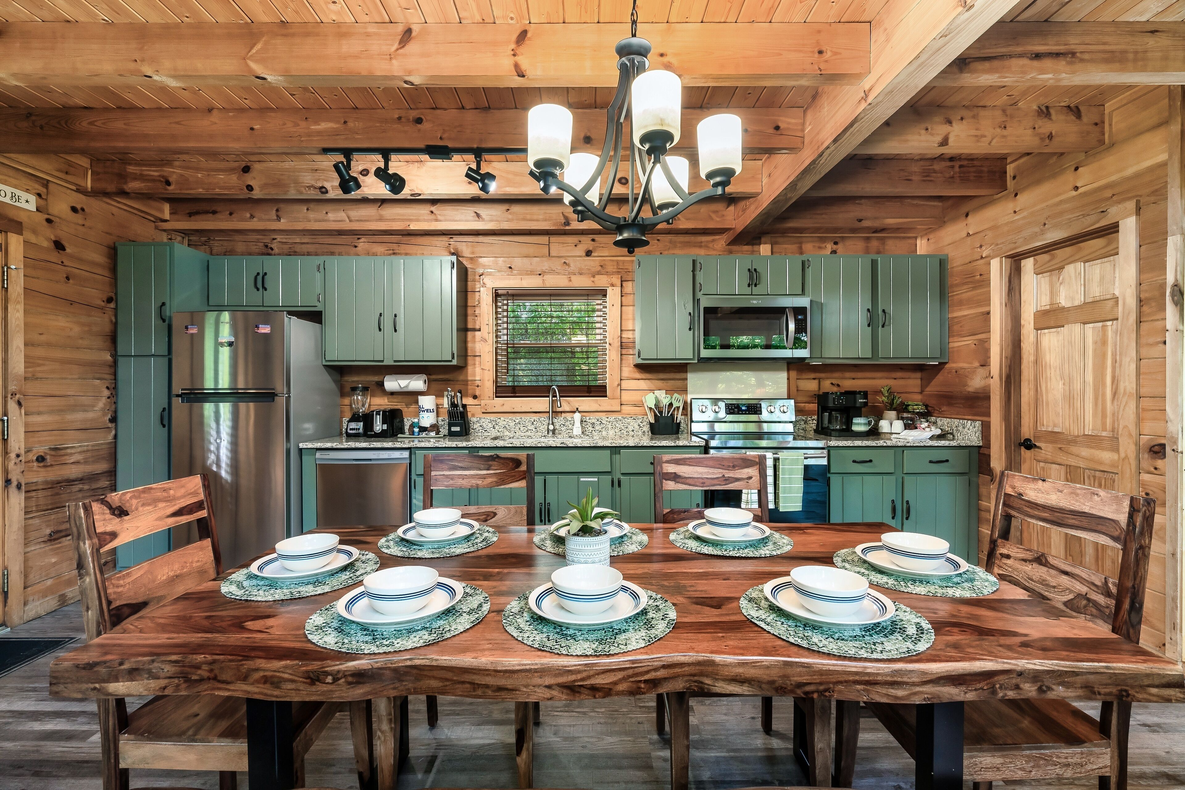 Une cuisine de style rustique, avec des meubles en bois, des armoires vertes, un réfrigérateur en acier inoxydable et une table à manger agrémentée d’assiettes et d’un vase.