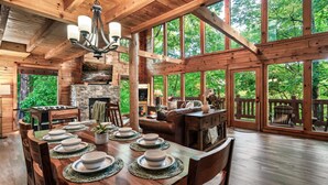A spacious wooden cabin with a dining area set for six, a stone fireplace, and large windows offering a view of the forest. - 15 Min. to Dollywood · Sauna · Hot Tub · Pool Table · Arcade • Foosball • Grill (sevierville)