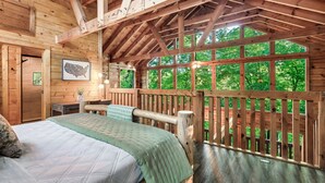 A wooden cabin room with a bed, a desk, and a view of the forest through large windows. - 15 Min. to Dollywood · Sauna · Hot Tub · Pool Table · Arcade • Foosball • Grill (sevierville)