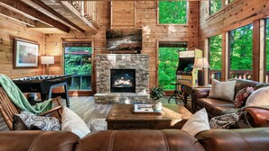 A cozy cabin interior with a stone fireplace, wooden walls, and large windows offering a view of the forest. - 15 Min. to Dollywood · Sauna · Hot Tub · Pool Table · Arcade • Foosball • Grill (sevierville)