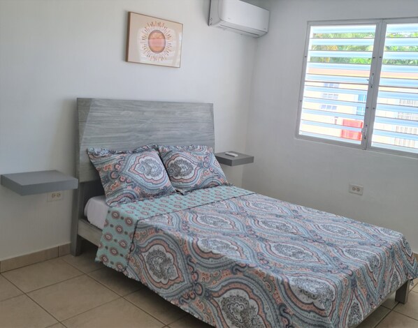 Une chambre à coucher avec un lit, une literie à motifs, une table de chevet, un climatiseur fixé au mur, une fenêtre avec des stores et un tableau encadré accroché au mur.