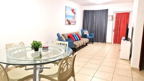 Una sala de estar con una mesa de vidrio, sillas color beige, un sofá con cojines de colores, un cuadro en la pared y un televisor sobre un mueble.