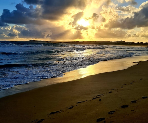 A beach with footprints leading to the water, waves, and a sunset in the background.