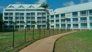 A multi-story hotel with a central courtyard, a pathway, and a fenced area with palm trees. - Charming studio with superb sea view (Le Gosier)