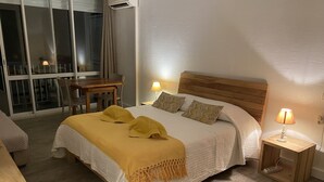 A hotel room with a bed, two bedside lamps, a wooden headboard, a window with curtains, and a table with chairs. - Charming studio with superb sea view (Le Gosier)