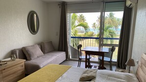 A hotel room with a bed, a sofa, a table, and a balcony with a view of palm trees and the sea. - Charming studio with superb sea view (Le Gosier)
