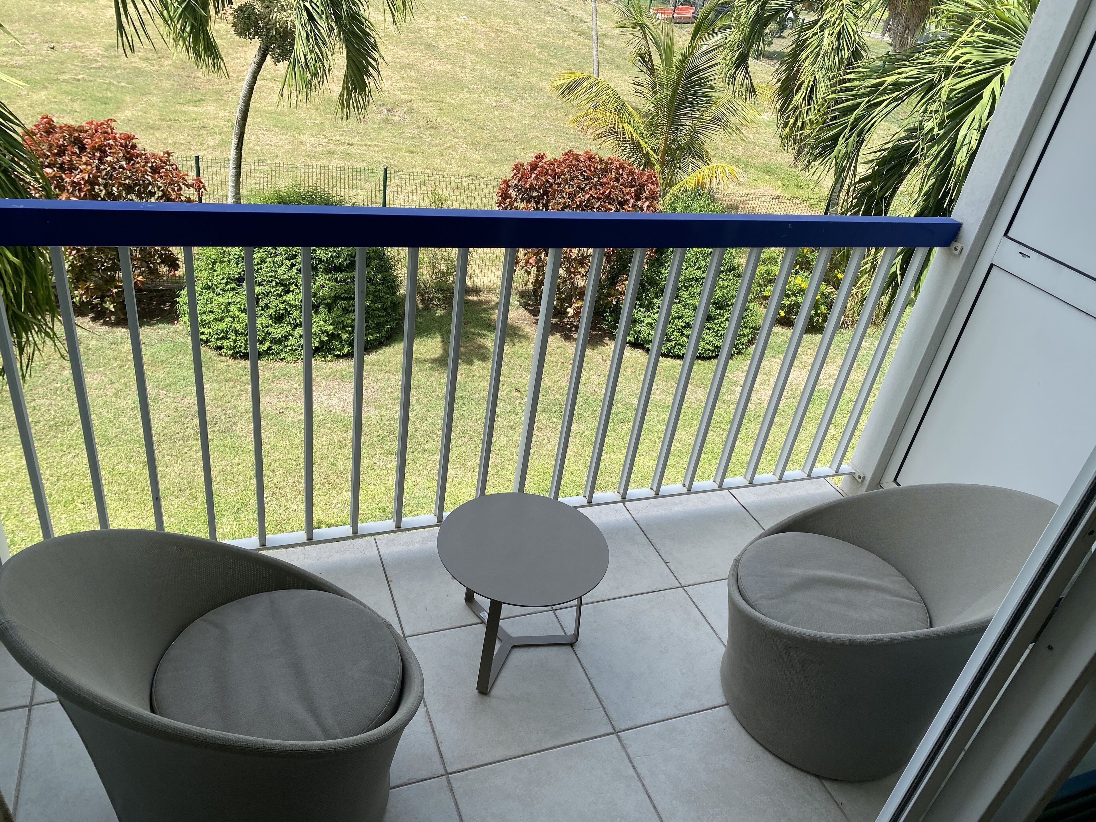 A balcony with two grey cushioned chairs and a small table, overlooking a well-maintained garden with palm trees and bushes.
