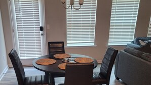 Un coin repas avec une table ronde, quatre chaises et un lustre. Il y a trois fenêtres équipées de stores, un canapé gris et un plancher en bois.
