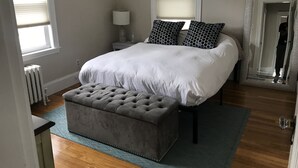 A bedroom with a large bed, a grey ottoman, a bedside table with a lamp, and a mirror. - Casa de Los Clarey • Boston • Logan • Beaches • Free Parking (Winthrop)