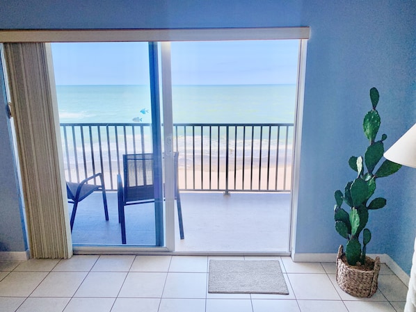 A room with a view of the beach through a sliding glass door, a chair, a potted plant, and a tiled floor.