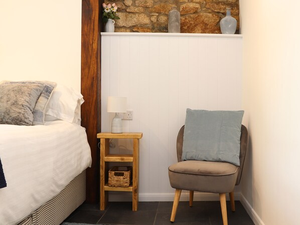 A bedroom with a bed, a nightstand with a lamp, a chair, and a stone wall. - The Dairy (Helston)