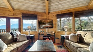 Ein gemütliches Blockhaus-Interieur mit Holzwänden, einem großen Fenster mit Blick auf die Berge, einer Couch, einem Couchtisch und einem an der Wand befestigten Fernseher.