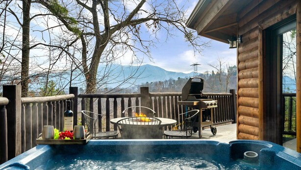 Ein Whirlpool mit Feuerstelle, umgeben von einer Holzdielen-Terrasse mit Blick auf eine gebirgige Landschaft.