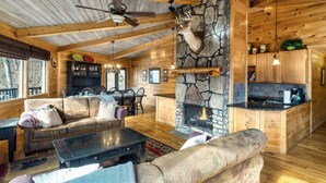 Ein gemütliches Blockhaus-Interieur mit Kamin aus Stein, Holzbalken, einem Essbereich und einem Wohnzimmer mit Sofas und Couchtisch.