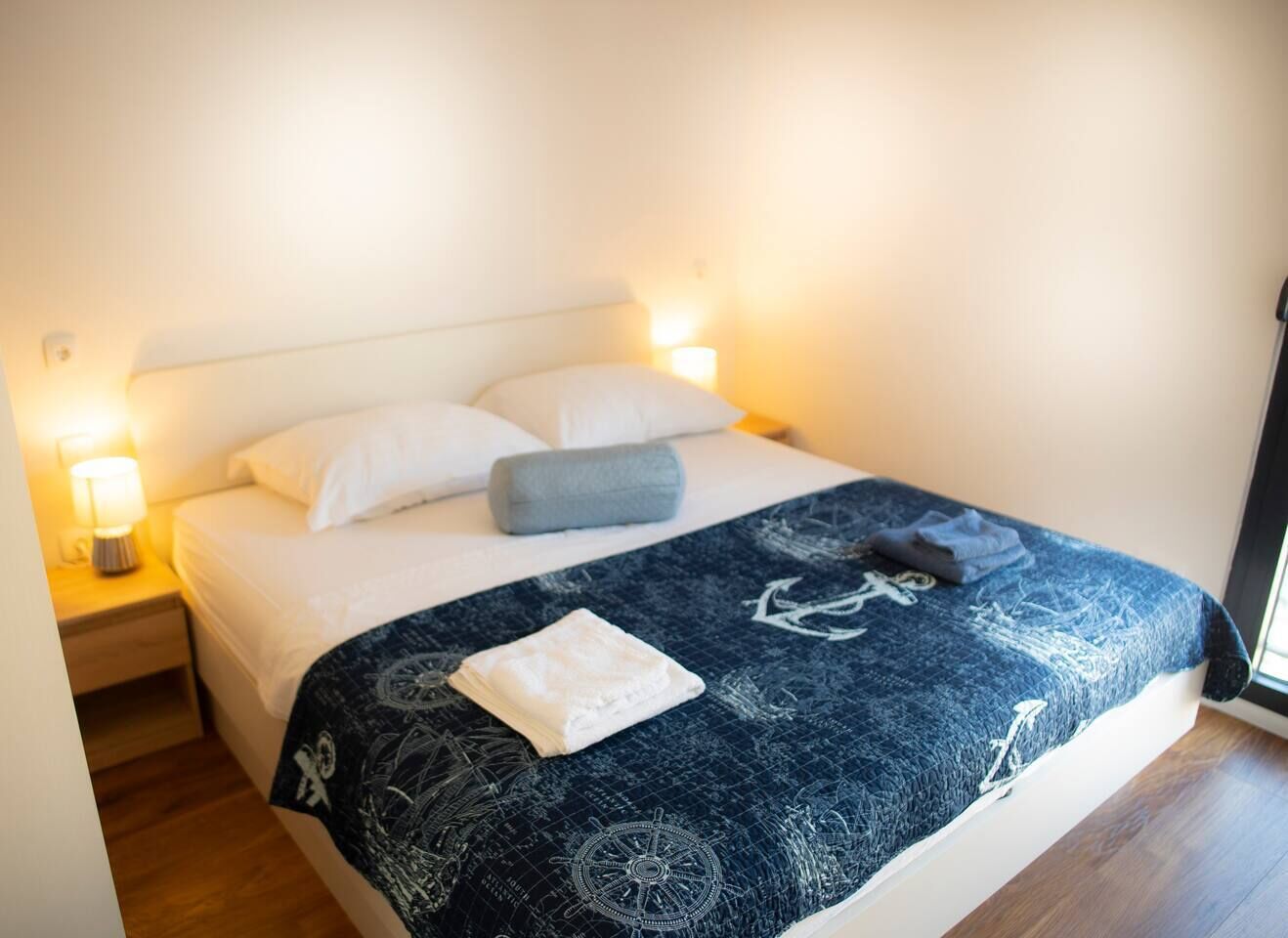 A neatly made bed with a patterned blanket, pillows, and a folded towel. There is a bedside lamp on a wooden nightstand.
