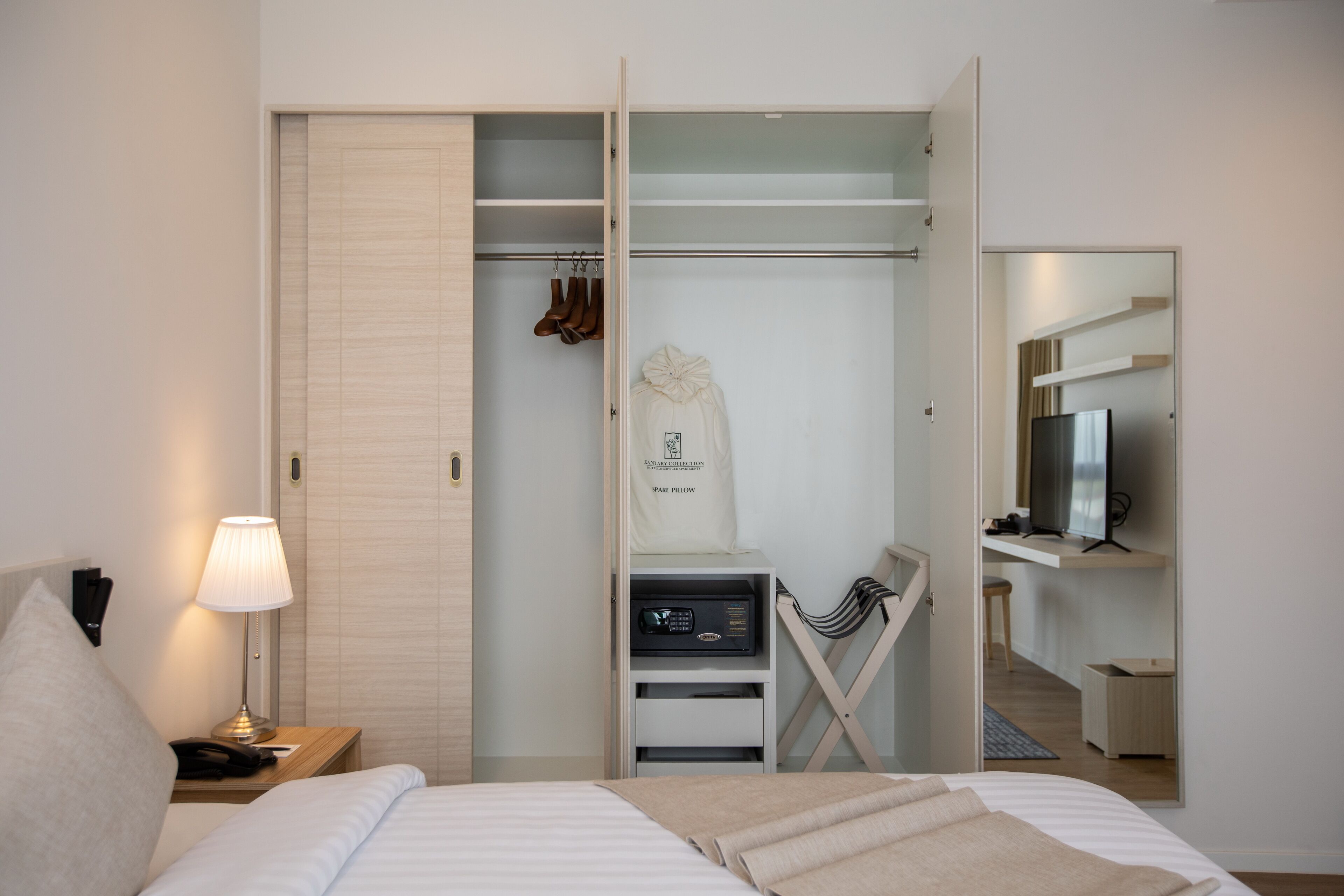 A hotel room with a bed, a bedside lamp, a wardrobe with hanging shoes, a coat, a desk with a computer, and a TV.