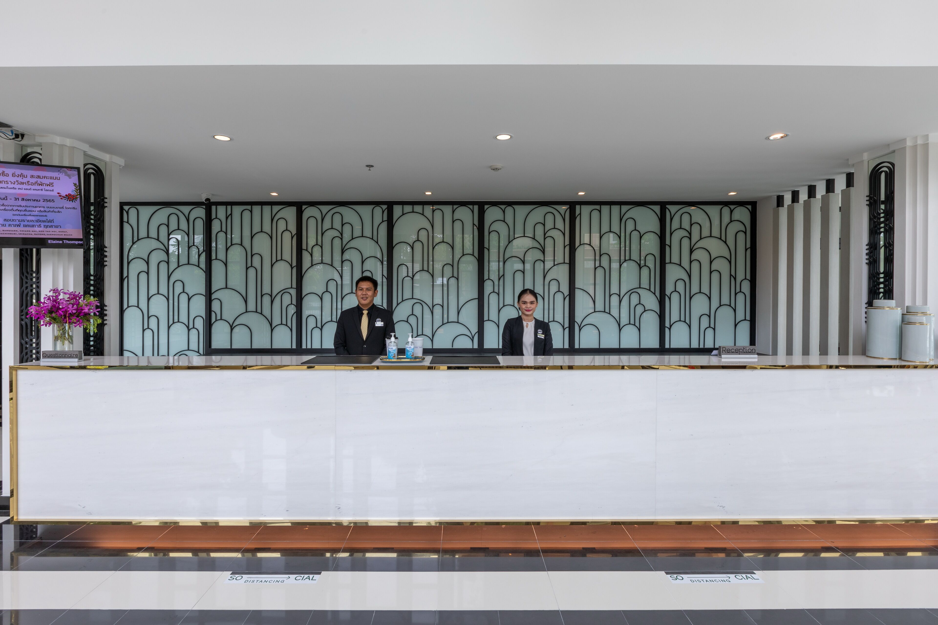 A reception desk with two staff members, a digital screen, and a decorative wall.