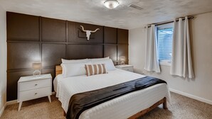 Een slaapkamer met een groot bed, een hoofdbord met een kopskelet van een koe, een nachtkastje met een lamp en een raam met gordijnen.