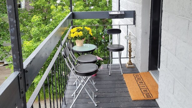 A balcony with a wooden floor, black metal railing, and a small table with two chairs. There are hanging flower pots with red and white flowers. - Vibrant apartment in the heart of the town (Edmundston)