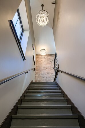 A staircase with a brick wall at the bottom, metal handrails, and a chandelier at the top. - Luxury Lofts, near UNL, Arena, Stadium, prime location #203  (Lincoln)