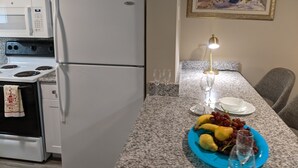 A kitchen with white appliances, a granite countertop, a dining table with a blue plate of fruit, and a framed floral painting on the wall. - Blissful Villa, King Bed (Hilton Head Island)