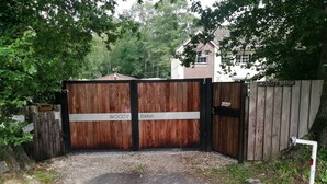 Een houten poort met de aanduidingen 'WOODY BANK' en 'WACHTHOND', die uitkomt op een grindpad.