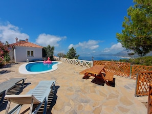 A pool with lounge chairs and a wooden table, surrounded by stone flooring and a white fence.