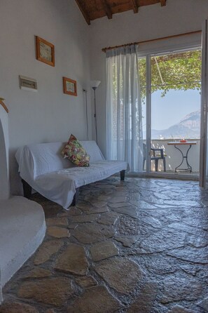 A stone-floored room with a white sofa, a colorful pillow, and a table with chairs outside a glass door. - Apartment With Pool and Panorama View (Samos)