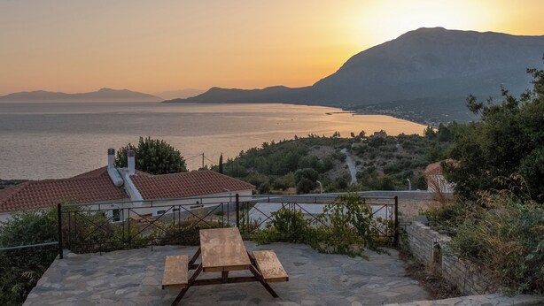 A stone patio with a wooden picnic table, overlooking a body of water and distant mountains during sunset.