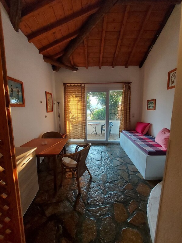 Une pièce dotée d’un sol en pierre, d’un plafond en bois, d’une table à manger avec des chaises, d’un canapé avec des coussins et d’une porte-fenêtre coulissante donnant sur un espace extérieur.