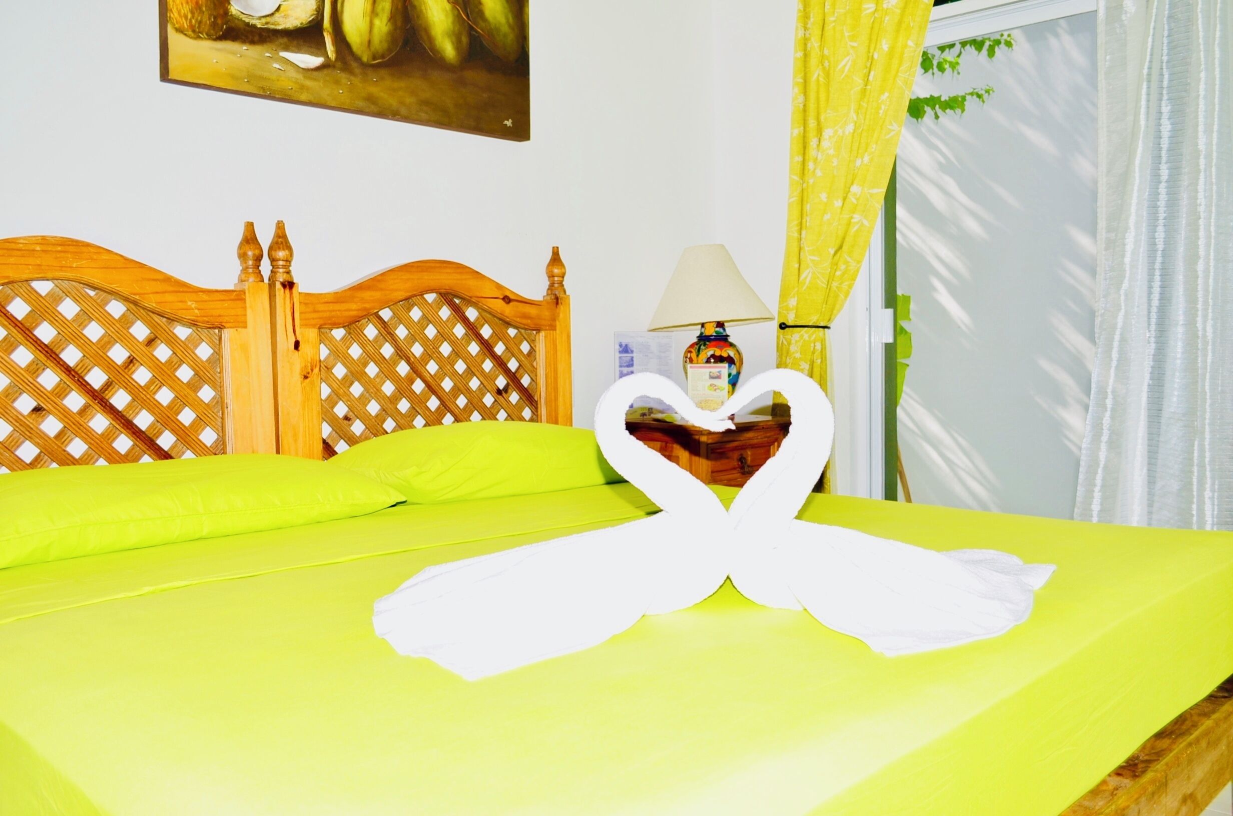 A bed with a bright green bedspread, a wooden headboard with a lattice design, a white towel folded in a heart shape, a lamp on a bedside table, and a painting on the wall.