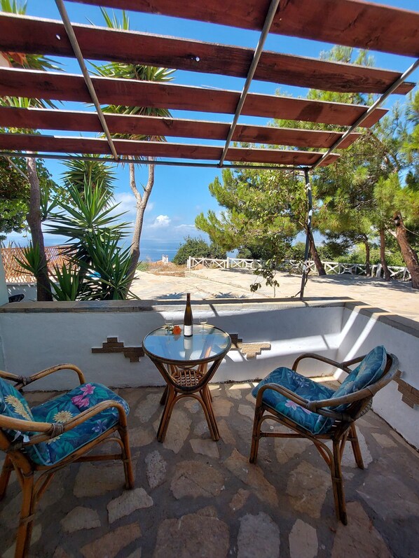 A covered outdoor seating area with a table and chairs, a bottle of wine, and a view of the sea.
