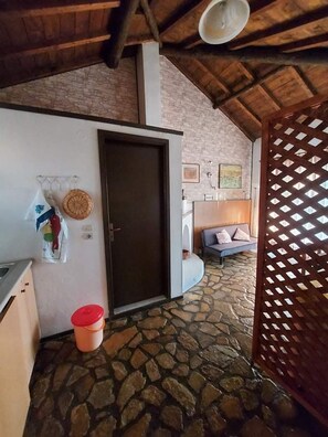 A room with a stone floor, a wooden ceiling, a door, a wall-mounted basket, and a small orange bucket.