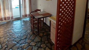 A room with a stone floor, wooden ceiling beams, a dining table with chairs, a kitchen area, and a large window with curtains. - Apartment 800m From Koumeika, Samos (Samos)