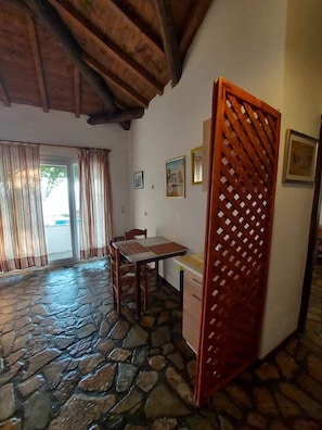 A room with a stone floor, wooden ceiling beams, a dining table with chairs, a kitchen area, and a large window with curtains. - Apartment 800m From Koumeika, Samos (Samos)