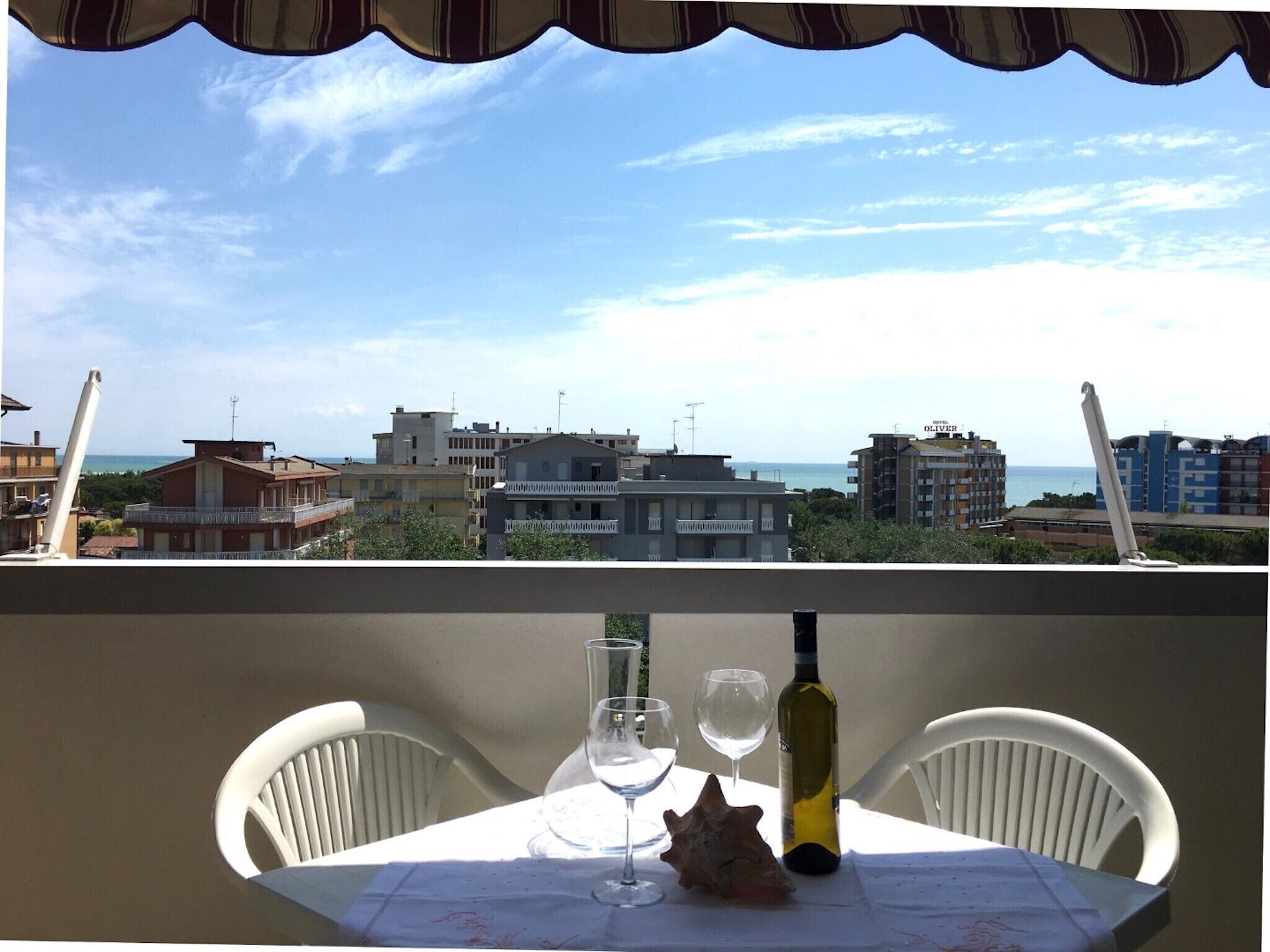 Une table avec une bouteille de vin, deux verres et un coquillage décoratif, sur un balcon donnant sur un paysage urbain avec des bâtiments et un ciel dégagé.