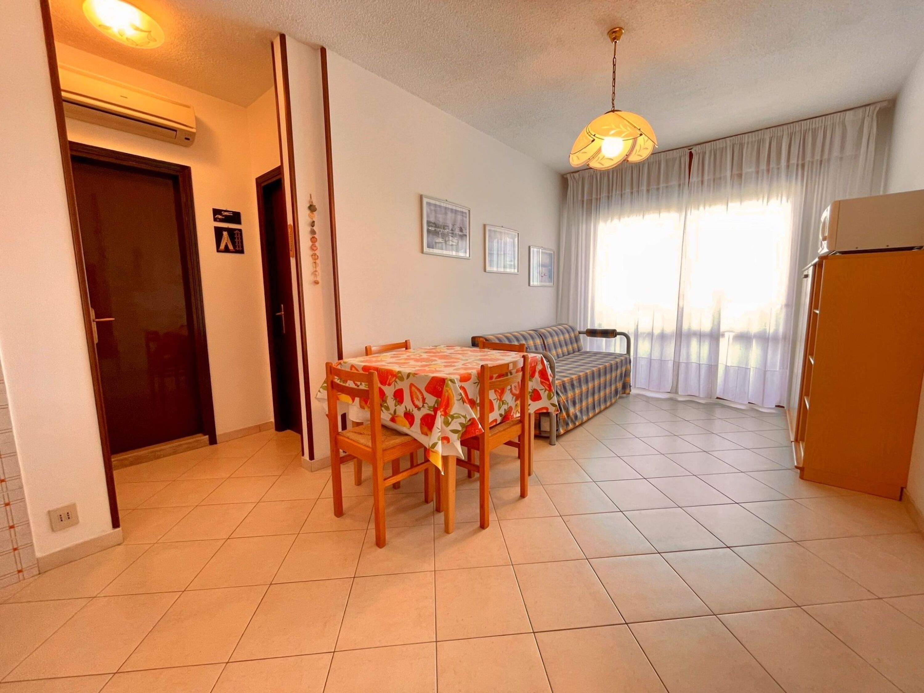 A compact living space with a dining table, chairs, a sofa, a ceiling light, and a window with curtains.
