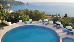 Ein Swimmingpool mit blauen Liegestühlen und Sonnenschirmen, umgeben von Grün, mit Blick auf eine Küstenlandschaft und das Meer mit Klippen.