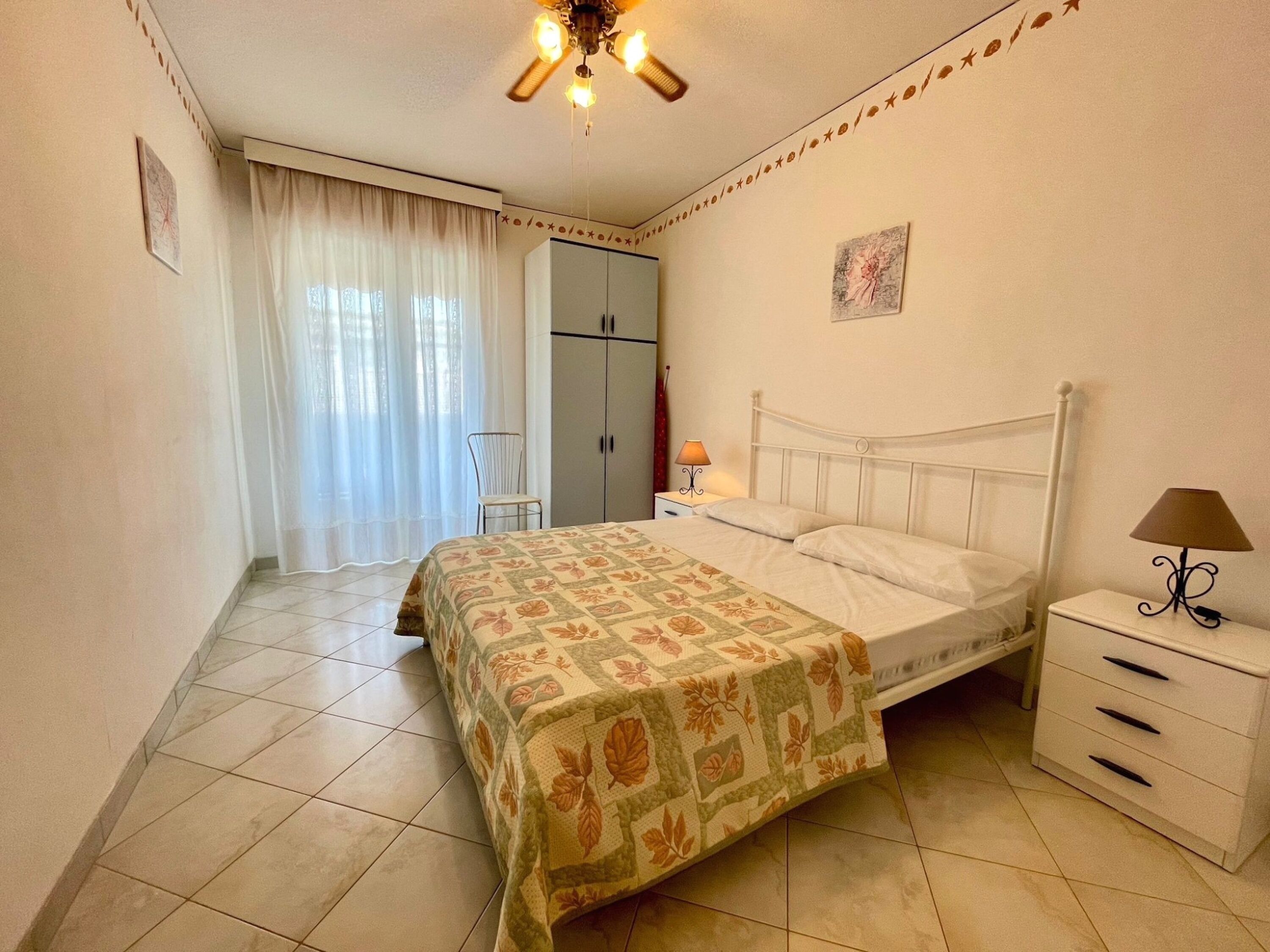 Quarto com cama, mesa de cabeceira, guarda-roupa, cadeira e ventilador de teto.