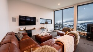 A modern living room with leather sofas, a flat-screen TV, and a large window offering a snowy mountain view.