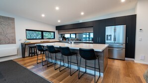 A modern kitchen with a central island, bar stools, a stainless steel refrigerator, and a large window providing a view of the outdoors.