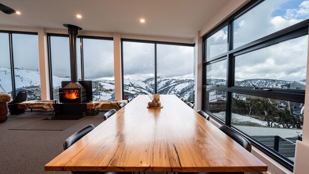A spacious room with a large wooden table, a fireplace, and large windows offering a snowy mountain view.
