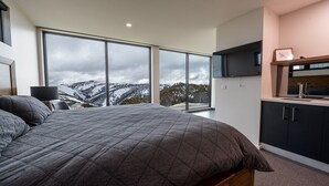 A bedroom with a large bed, a TV mounted above the kitchen counter, and a view of snow-covered mountains through large windows.