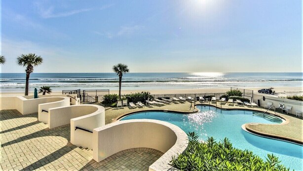 Ein Poolbereich mit Blick auf Strand und Meer, umgeben von Liegestühlen und Palmen.